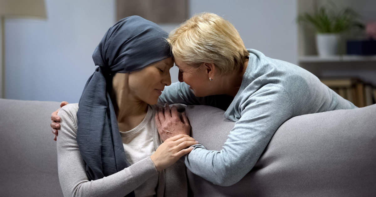 A health care proxy, holding hands with a woman with a scarf around her head sitting on the couch